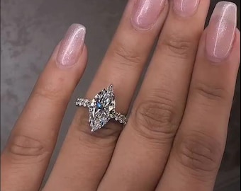 May include: A marquise-cut diamond engagement ring with a pavé band. The ring is set on a hand with long, light pink nails. The diamond is the focal point, reflecting light and set in a silver band.