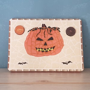 Peut inclure: Un plateau en céramique blanche avec un motif en mosaïque présente une citrouille d'Halloween avec un sourire narquois. Deux petits bougeoirs sont de chaque côté de la citrouille.