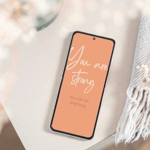 Könnte beinhalten: Ein Smartphone mit einem pfirsichfarbenen Bildschirm, auf dem der Text "You are strong. You can do anything." angezeigt wird.