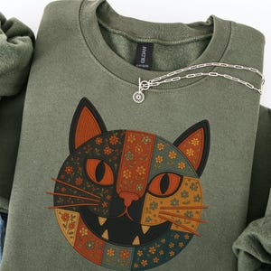 Könnte beinhalten: Ein olivgrünes Sweatshirt mit einem Katzen-Gesicht-Design. Das Katzengesicht ist geteilt, wobei eine Seite ein Patchwork aus orangefarbenen, braunen und blauen Blumenmustern aufweist. Eine silberne Halskette mit einem kreisförmigen Anhänger liegt auf dem Sweatshirt.