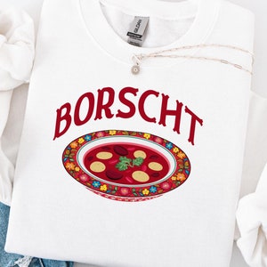 Peut inclure: Sweat-shirt blanc avec le mot "BORSCHT" en lettres rouges et une illustration colorée d'un bol de bortsch.