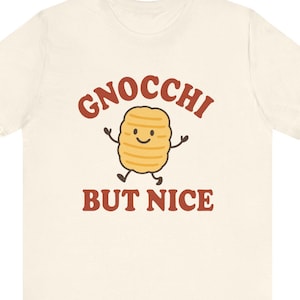 May include: Cream-colored t-shirt featuring a cartoon gnocchi character with arms and legs. The text "GNOCCHI BUT NICE" is printed above and below the character in a brown, rounded font. The gnocchi has a smiling face.