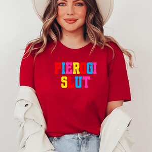 May include: A red t-shirt with the text "PIEROGI SLUT" in colorful block letters. The person in the image is wearing a cream-colored hat, a white jacket, and blue jeans.