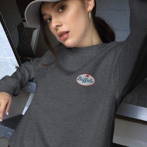 Könnte beinhalten: Ein dunkelgraues Sweatshirt mit einem rot-weiß-blauen ovalen Aufnäher, auf dem "Buffalo" steht. Das Sweatshirt wird von einer Person mit einer grauen Baseballkappe getragen.