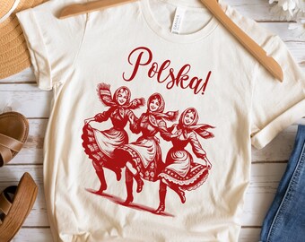 以下が含まれることがあります： クリーム色のTシャツ。赤い筆記体で「Polska！」の文字が書かれ、その上に伝統的なポーランド民族舞踊の衣装を着た3人の女性の赤いイラストが描かれています。木製のハンガーにかけられています。