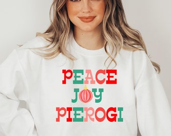 Może przedstawiać: Biała bluza z czerwono-zielono-różowym nadrukiem z napisem "Peace Joy Pierogi" z różową ozdobą między słowami "Joy" i "Pierogi".