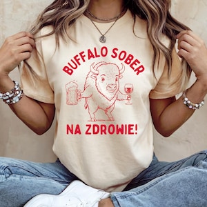 Puede incluir: Camiseta color crema con un gráfico rojo de un búfalo sosteniendo una jarra de cerveza y una copa de vino. El texto "BUFFALO SOBER" está arqueado sobre el búfalo, y "NA ZDROWIE!" está impreso debajo.