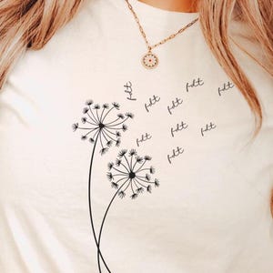 May include: Cream-colored t-shirt featuring a black dandelion design with the word "felt" repeated. The dandelion illustration includes two flower heads with delicate stems. The shirt is made of a soft material.