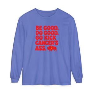 May include: A periwinkle blue long-sleeve shirt featuring red text that reads BE GOOD. DO GOOD. GO KICK CANCER'S ASS. Below the text is a red silhouette of a buffalo with an orange awareness ribbon integrated into its body design.