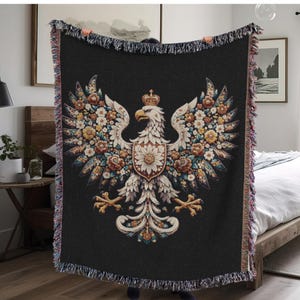 May include: A black woven throw blanket with a white eagle design. The eagle is surrounded by floral details and has a crown on its head.