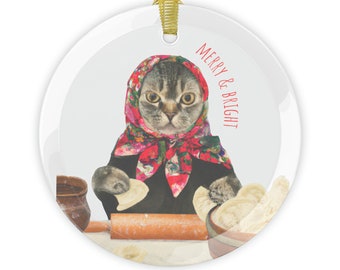 May include: A round glass ornament with a cat wearing a red and white headscarf and black clothing. The cat is making dumplings. The text "Merry & Bright" is written in red on the ornament.