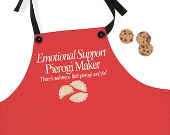May include: A red apron with black ties and buttons. The apron has the text "Emotional Support Pierogi Maker" and "There's nothing a little pierogi can't fix!" with a pierogi graphic. Three chocolate chip cookies are next to the apron.