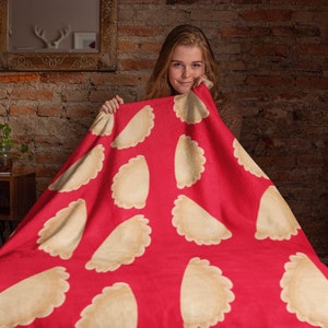 May include: A red blanket with a repeating pattern of pierogi dumplings. The dumplings are beige with a scalloped edge, set against a vibrant red background. The blanket is held up by a person.