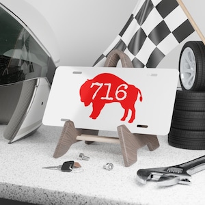 May include: White metal license plate with a red buffalo silhouette and the number 716 printed on it. The plate is on a wooden stand.