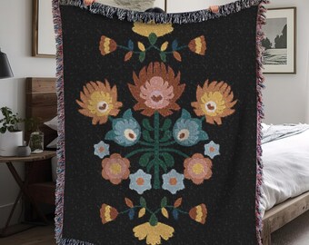 Puede incluir: Una manta de tejido negro con un patrón floral en tonos de rosa, amarillo, azul y verde. Las flores están dispuestas en un diseño simétrico con un tallo central y hojas.