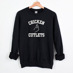 May include: Black sweatshirt with white text that reads "CHICKEN CUTLETS" with a hand gesture above the word "CUTLETS".