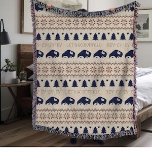May include: A beige woven throw blanket with a repeating pattern of dark blue buffalo, snowflakes, and evergreen trees. The text "LET'S GO BUFFALO" and "HEY-EY-EY-E" are also displayed. The blanket has a fringed edge.