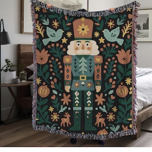 May include: A black woven throw blanket featuring a colorful design of a nutcracker soldier, birds, flowers, and deer. The blanket has a fringed edge and is held up by a person.