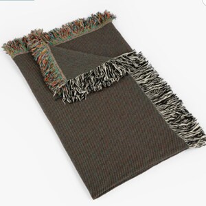 May include: A folded brown blanket with fringed edges. The fringe on one side is a mix of colors, while the other side has black and white fringe. The blanket is made of a woven material.