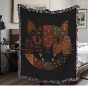 May include: Black woven throw blanket with a cat face design. The cat's face is divided into sections with floral and geometric patterns in brown, orange, and green. The blanket has a fringed edge. Measures approximately 150cm x 200cm.