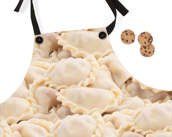 May include: An apron featuring a close-up image of pierogies, a traditional Eastern European dumpling. The apron has black ties and buttons. Three chocolate chip cookies are visible on the right side of the image. The apron is designed for cooking and baking.