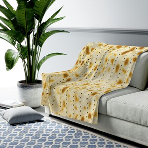 May include: A soft, plush throw blanket with a pattern of matzah, a traditional unleavened bread eaten during Passover. The blanket is draped over a light gray couch with a green plant in a pot in the background.