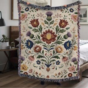 May include: Cream-coloured woven throw blanket with a floral pattern in shades of red, blue, yellow, and purple. The design features stylised flowers and leaves. The blanket has a fringed edge and is held up by a person.