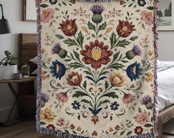 Peut inclure: Plaid tissé de couleur crème avec un motif floral dans les tons de rouge, bleu, jaune et violet. Le motif présente des fleurs et des feuilles stylisées. La couverture a un bord à franges et est tenue par une personne.