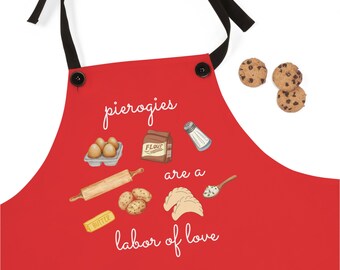 May include: Red kitchen apron with a white design that says "pierogies are a labor of love". The design includes illustrations of ingredients for making pierogies, such as eggs, flour, butter, and a rolling pin.
