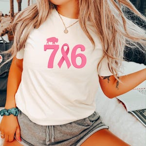 Op de afbeelding: Crèmekleurig T-shirt met een roze afbeelding. De afbeelding bevat een buffelsilhouet boven het nummer 786, waarbij het nummer 8 is vervangen door een roze lint. Het shirt heeft korte mouwen.