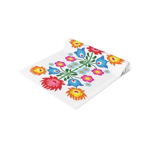 May include: A white table runner with a colorful floral design. The design features red, pink, yellow, orange, blue, and green flowers. The flowers are arranged in a symmetrical pattern.