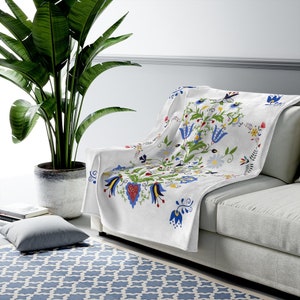May include: White fleece throw blanket with a colorful floral pattern. The pattern features blue, red, yellow, and green flowers and leaves. The blanket is folded and draped over a white couch.