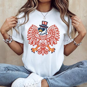 May include: White t-shirt featuring a patriotic eagle design. The eagle is depicted with red and white wings, wearing sunglasses and a top hat with stars. The design is centered on the shirt.
