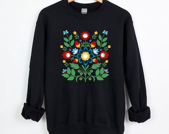 Puede incluir: Sudadera negra con un vibrante diseño floral. El diseño incluye flores y hojas rojas, azules, amarillas y verdes dispuestas simétricamente. La sudadera está colgada en una percha de madera.