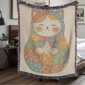 Op de afbeelding: Een beige sprei met een lachend kattenontwerp. De kat draagt een muts en een patchwork-outfit in pastelkleuren. De deken heeft een gefranjerde rand en een grillige, cartoonachtige stijl.