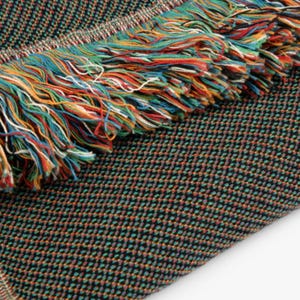 May include: A black and green woven throw blanket with a colorful fringe. The fringe is made of yarn in shades of red, orange, yellow, green, blue, and white.