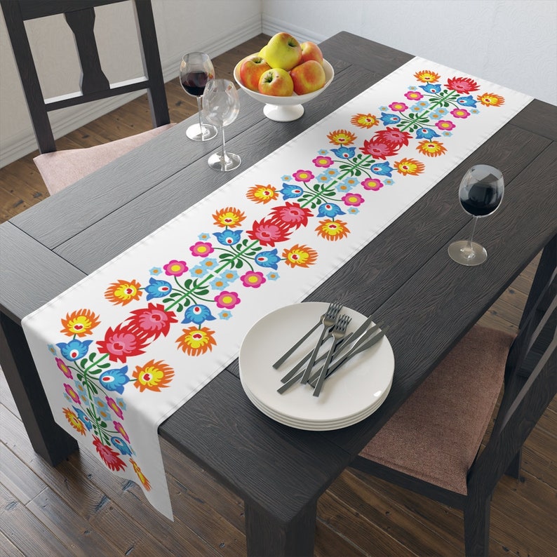May include: White table runner with a colorful floral pattern. The pattern features red, blue, yellow, and pink flowers with green stems. The runner is on a dark wood table with a white plate and silverware.