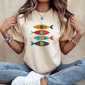 May include: Cream-colored t-shirt featuring a design of four stylized fish. Each fish is a different color: yellow, red, blue, and teal, and decorated with floral patterns. The shirt is a casual, everyday wear item.
