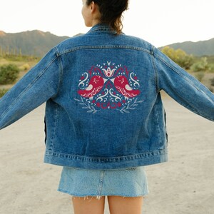 May include: A blue denim jacket with a white and red embroidered design of two birds facing each other. The design features floral elements and a crown.