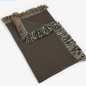 May include: A green and brown woven throw blanket with a black and white fringe border. The blanket has a textured weave and is folded in half.