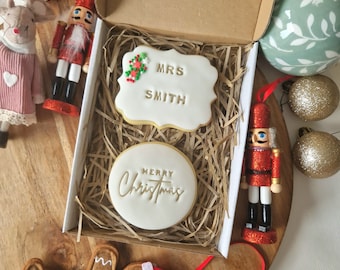 May include: A gift box containing two decorated Christmas cookies. One cookie is rectangular with "MRS SMITH" and festive accents. The other is round and says "MERRY Christmas". The box is on a wooden surface with Christmas ornaments.