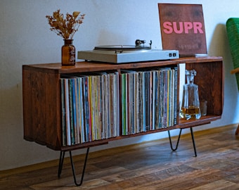 May include: A mid-century modern record player console with a collection of vinyl records. The wooden cabinet has black hairpin legs and a turntable on top. A green armchair and a brown sign with the word "SUPR" are in the background.