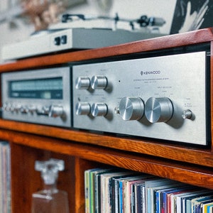 May include: A vintage silver Kenwood stereo receiver with a wooden cabinet and a record player on top. The receiver has a volume knob, input selector, and other controls. The cabinet has a shelf below the receiver that is filled with vinyl records.