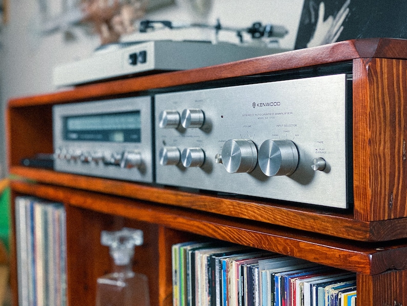 May include: A vintage silver Kenwood stereo receiver with a wooden cabinet and a record player on top. The receiver has a volume knob, input selector, and other controls. The cabinet has a shelf below the receiver that is filled with vinyl records.