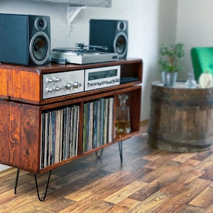 May include: A wooden record player stand with a record player, two speakers, and a shelf for vinyl records. The stand has a dark brown finish and is on black hairpin legs.