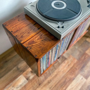 May include: A wooden record player stand with a turntable on top and shelves filled with vinyl records. The stand is stained a dark brown color and has a rustic look.