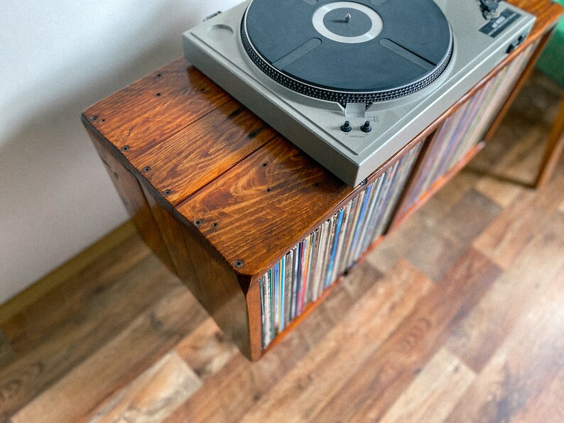 May include: A wooden record player stand with a turntable on top and shelves filled with vinyl records. The stand is stained a dark brown color and has a rustic look.