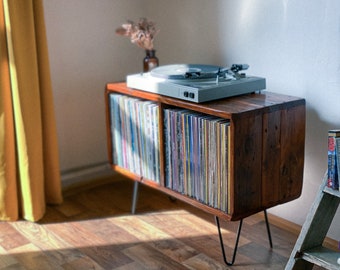 May include: A wooden record player stand with a turntable on top and two shelves filled with vinyl records. The stand has black hairpin legs and is sitting on a wooden floor.