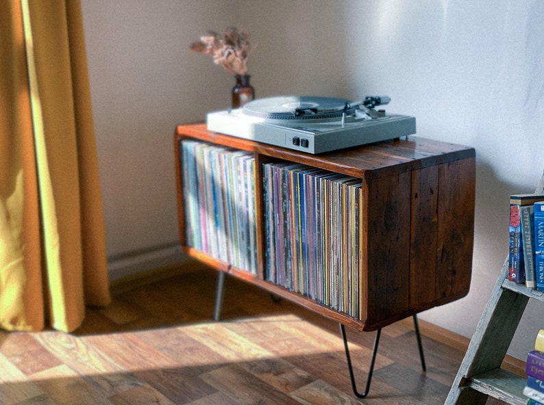 May include: A wooden record player stand with a turntable on top and two shelves filled with vinyl records. The stand has black hairpin legs and is sitting on a wooden floor.
