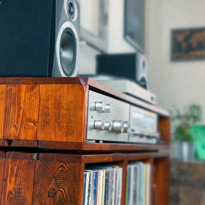 May include: A wooden record player stand with two shelves. The top shelf has a silver stereo receiver and two black speakers. The bottom shelf has a collection of vinyl records.